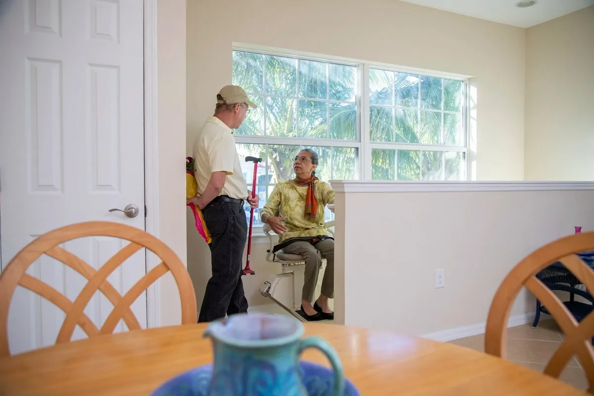 A man steadies an older woman holding a cane at the bottom of a staircase